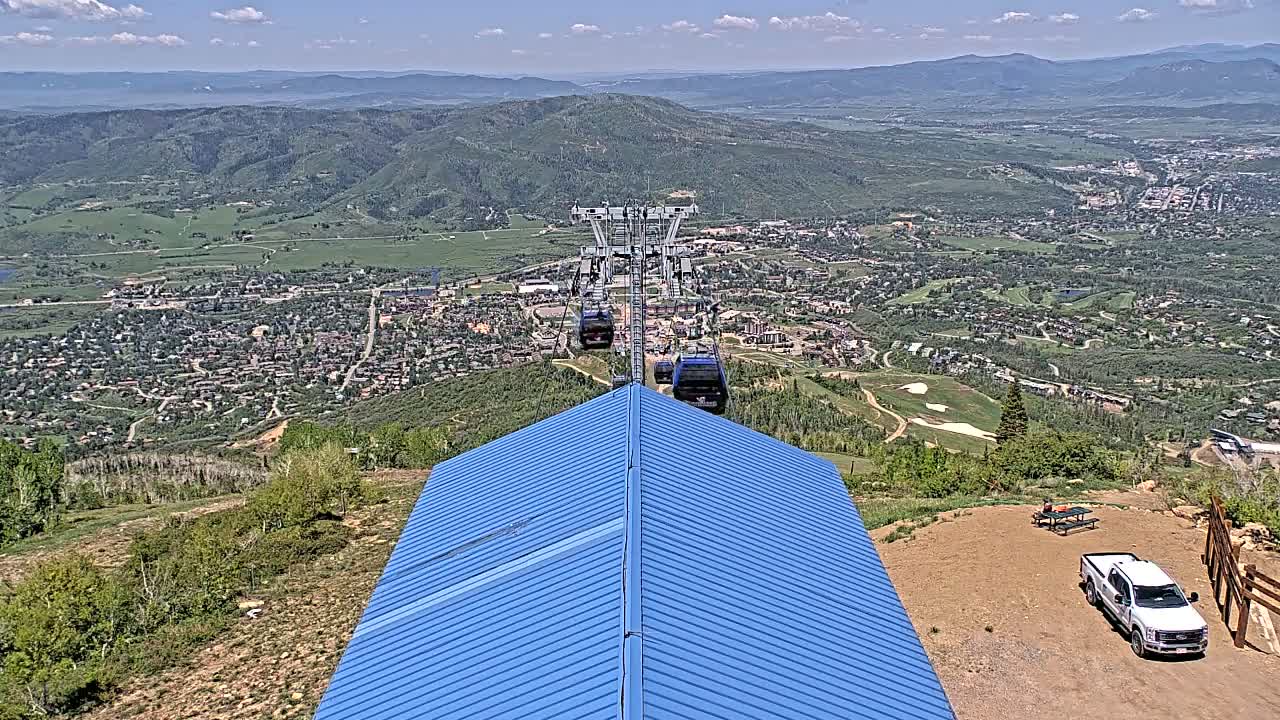 A blue-roofed building sits atop a mountain overlooking a valley town on a sunny day, with a chairlift ascending from the town below.