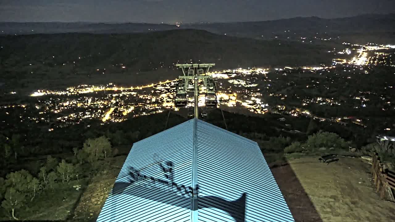 At night, a chairlift ascends a mountain overlooking a brightly lit town, with a building's roof in the foreground.