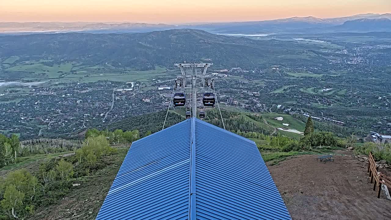 A mountaintop view at sunrise shows a chairlift ascending towards a distant valley town, with a blue metal roof in the foreground.