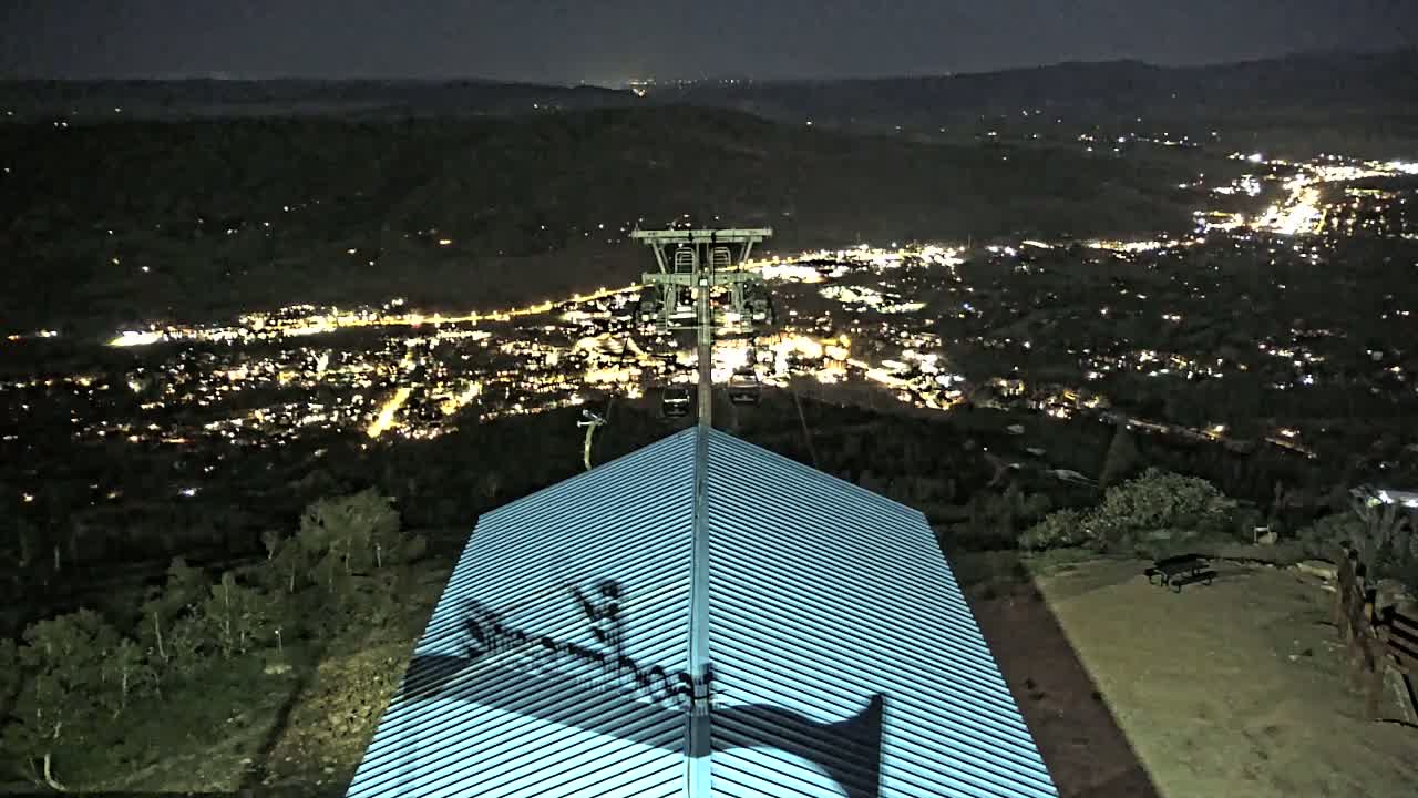 A nighttime view from a hilltop shows a city illuminated below, a chairlift in the middle ground, and a building's ridged roof in the foreground.