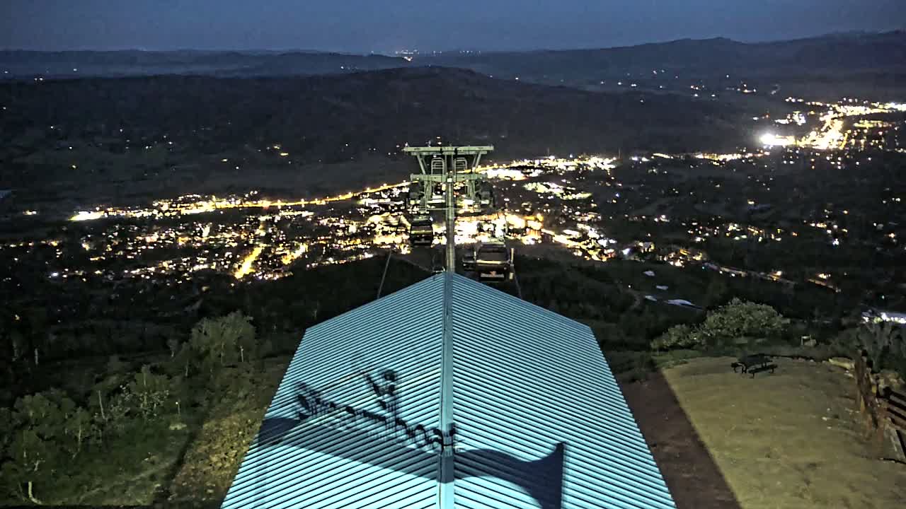 A nighttime view from a mountaintop shows a chairlift ascending towards a brightly lit town below, with the chairlift's shadow cast on a light teal roof.