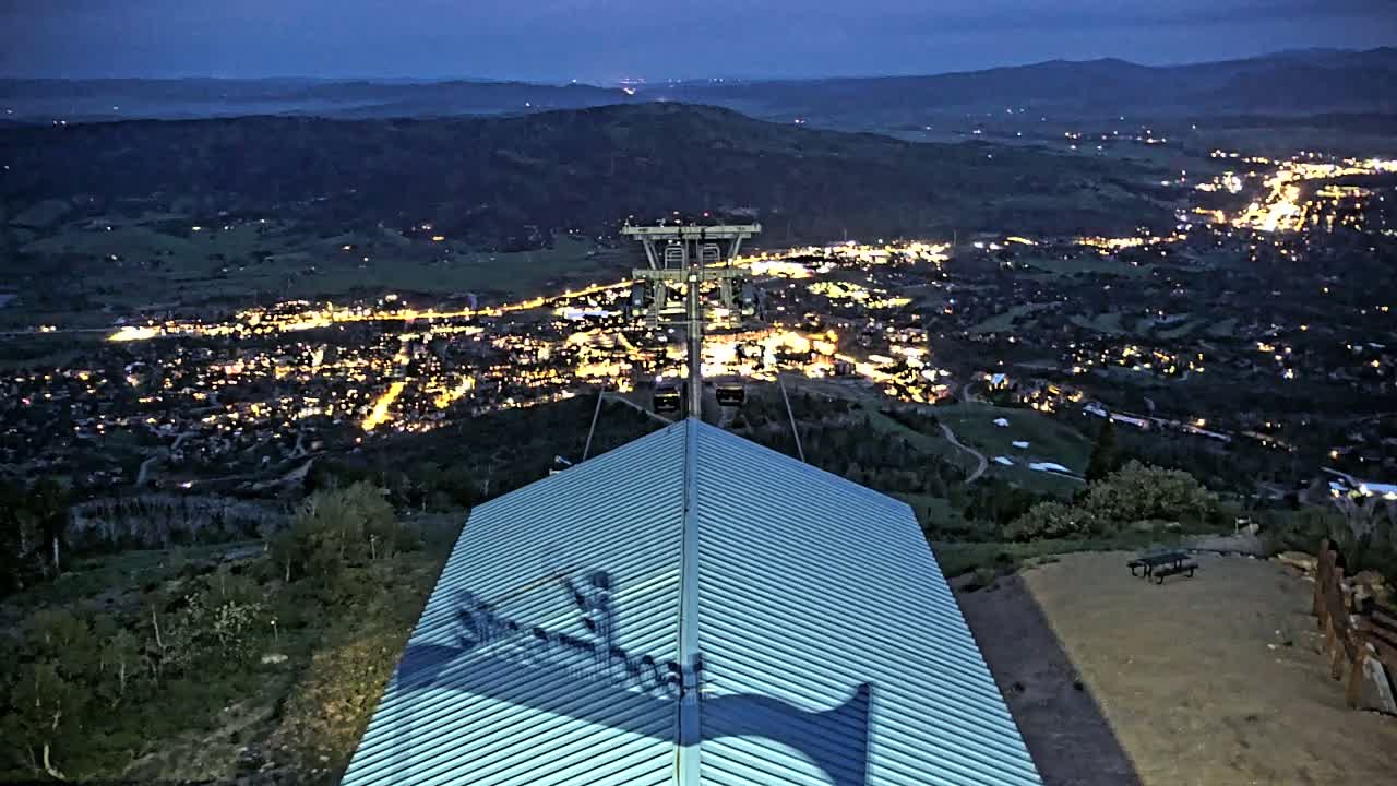 A metal roofed structure overlooks a valley town illuminated at night under a dark blue twilight sky.