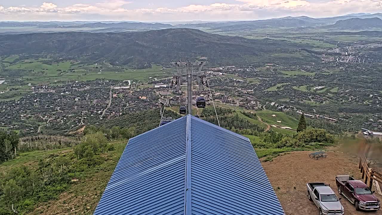 A blue-roofed structure overlooks a valley town under partly cloudy skies, with a chairlift ascending from the valley floor in the background.