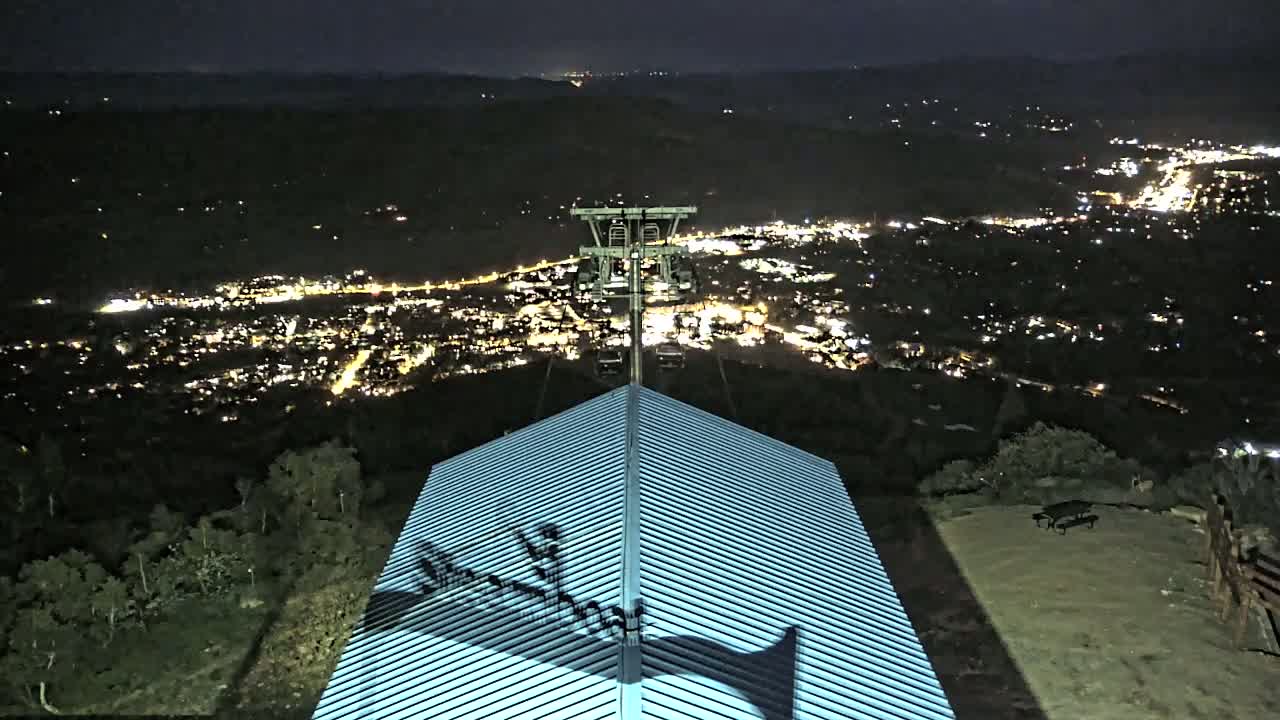 A nighttime view from a hilltop shows a city illuminated below, with a chairlift in the middle ground and a ridged roof in the foreground, under a clear, dark sky.