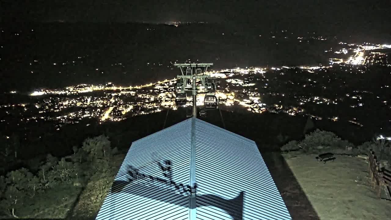 A nighttime view from a mountaintop shows a chairlift ascending toward a brightly lit city below, with a ridged roof in the foreground.