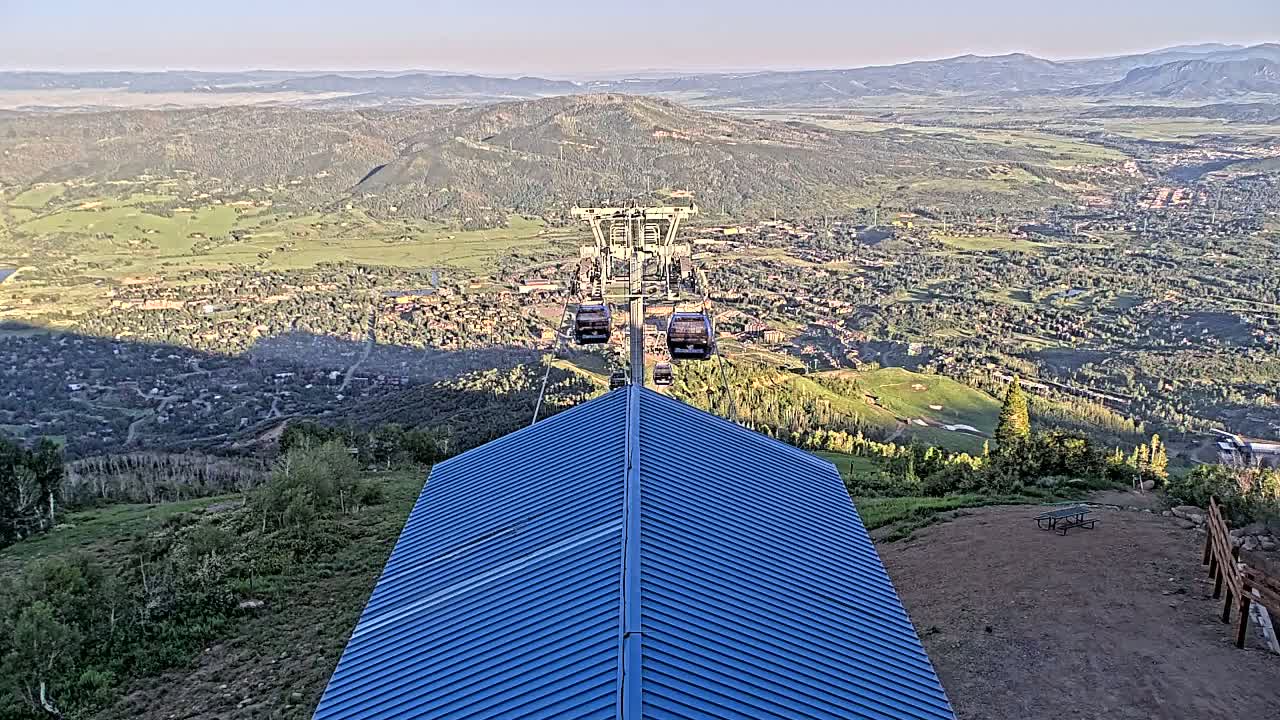 A blue-roofed structure overlooks a valley town and mountains under a clear sky, with a chairlift ascending from the town toward the structure.