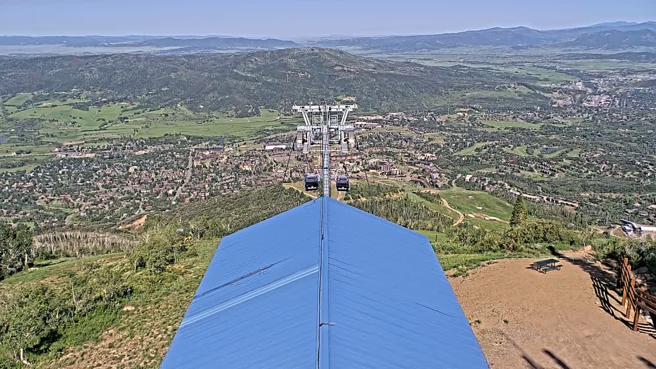 A chairlift ascends a mountain on a clear, sunny day, overlooking a valley town nestled amongst green hills and mountains in the distance.