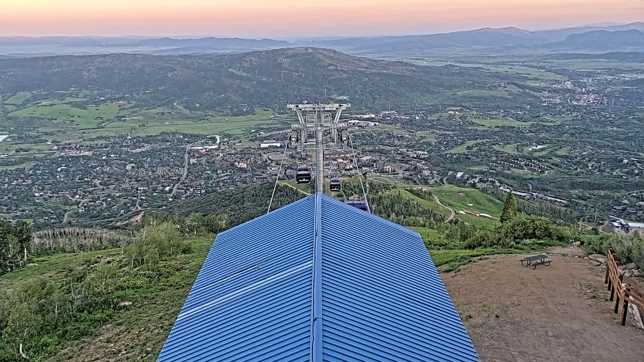 A chairlift ascends a mountain overlooking a valley town at sunset, with a blue roofed structure in the foreground.