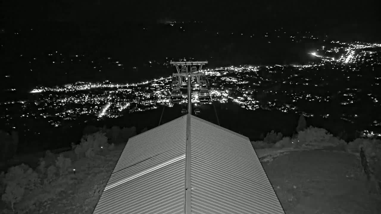 A nighttime view from a ridged roof shows a chairlift ascending towards a distant, brightly lit city under a clear sky.