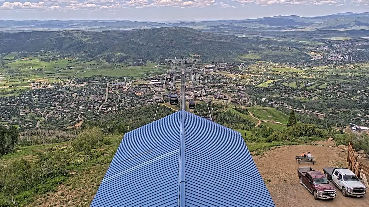 A blue-roofed structure sits atop a hill overlooking a valley town under partly cloudy skies, with a chairlift visible in the distance.