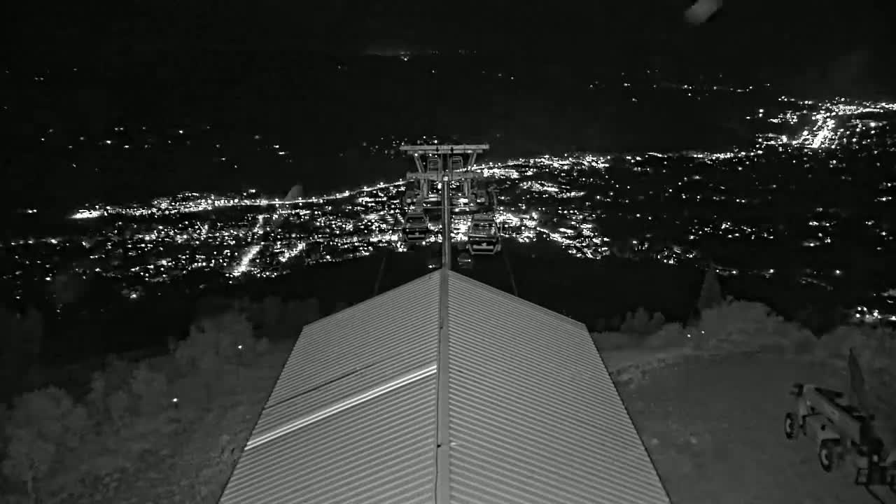 A nighttime view from a mountaintop shows a chairlift ascending towards a brightly lit city below.