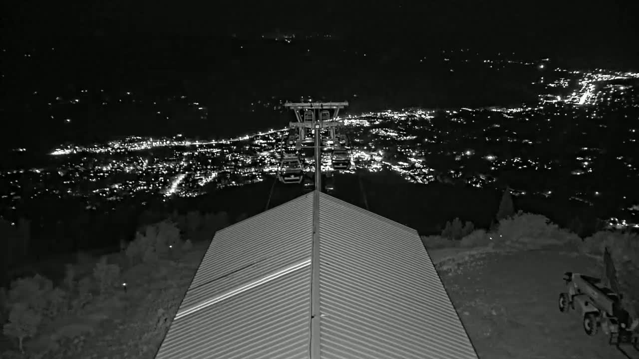 A nighttime view from a mountaintop shows a chairlift ascending toward a city glowing with lights below, as seen from a corrugated metal roof.