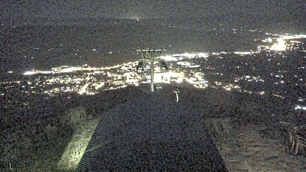 A nighttime view from a mountaintop shows a chairlift and a sprawling city illuminated below.