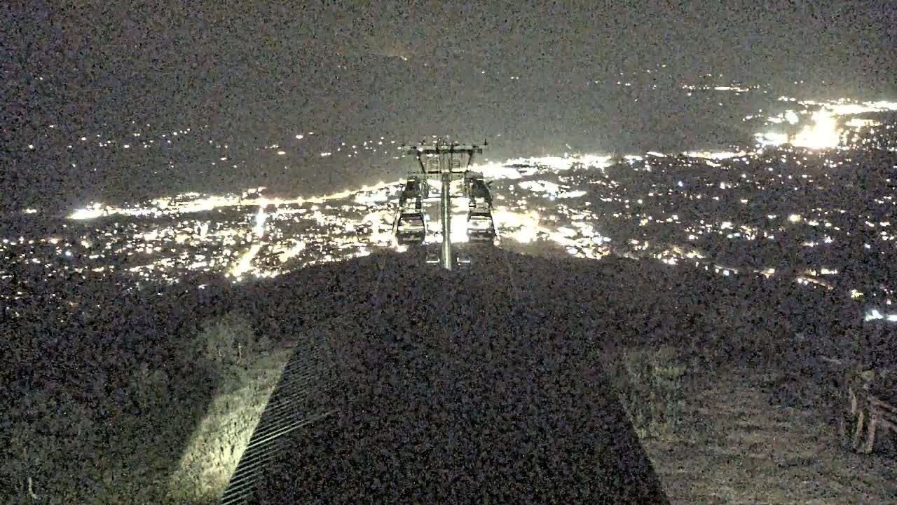 A nighttime view from a mountaintop shows a chairlift and a sprawling city illuminated below.