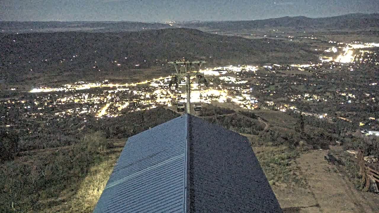 A nighttime view from a hilltop shows a brightly lit town below, a ski lift, and a two-toned roof in the foreground.