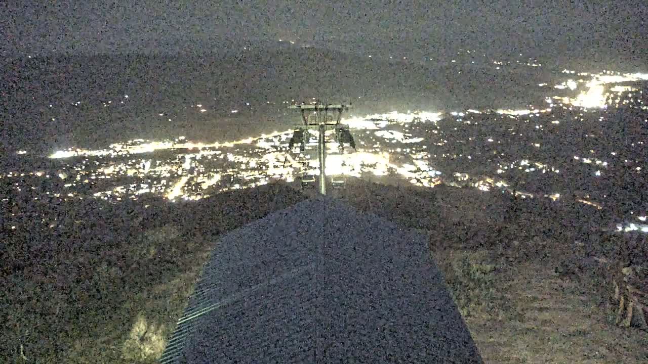 A nighttime view from a hilltop shows a brightly lit town below, a ski lift, and a two-toned roof in the foreground.