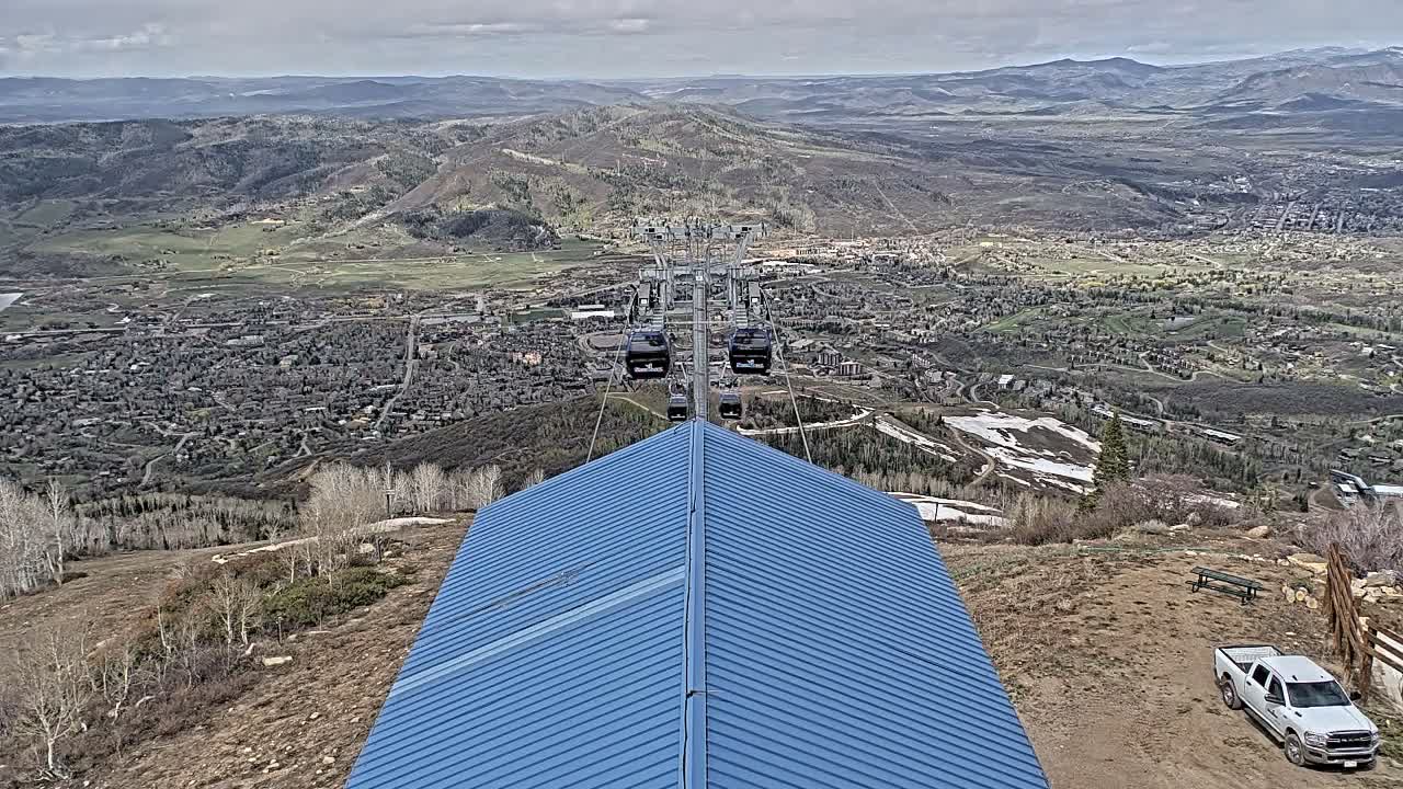 A night view from a snow-covered ski lift station looks out over a brightly lit, distant snow-dusted city under an overcast sky, with light snow falling or recently fallen.