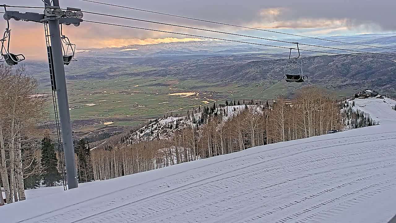 A snow-covered ski slope overlooks a valley under a cloudy sky with hints of sunset.