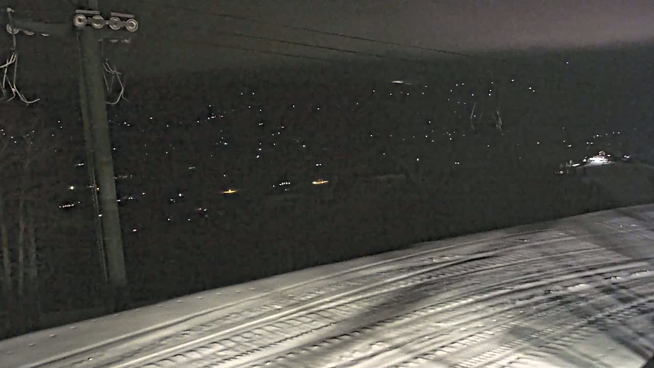 A snow-covered slope at night shows tire tracks leading toward a distant town with faint lights.