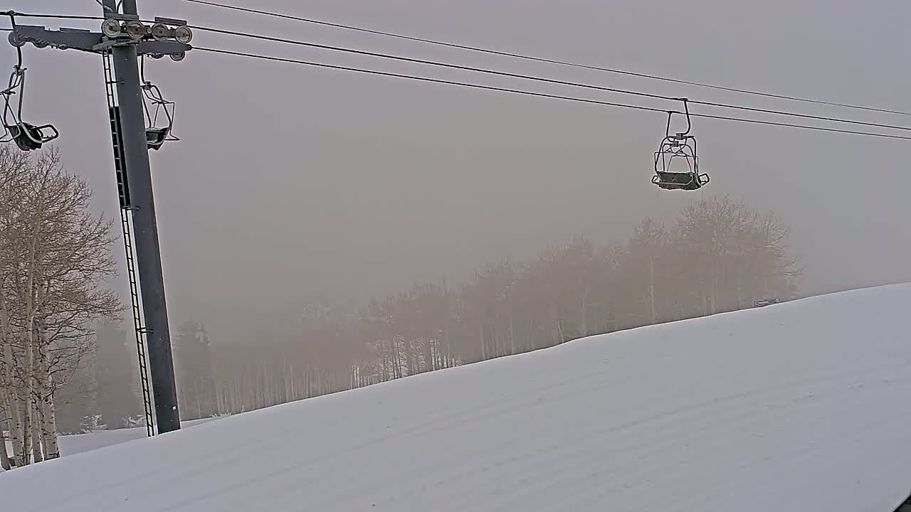 A ski lift chair hangs on a cable above a snowy slope with a hazy, overcast sky and bare trees in the background.