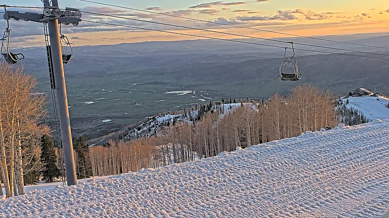 A snowy mountain slope at sunset, with a ski lift and empty chairlifts visible, overlooks a valley.