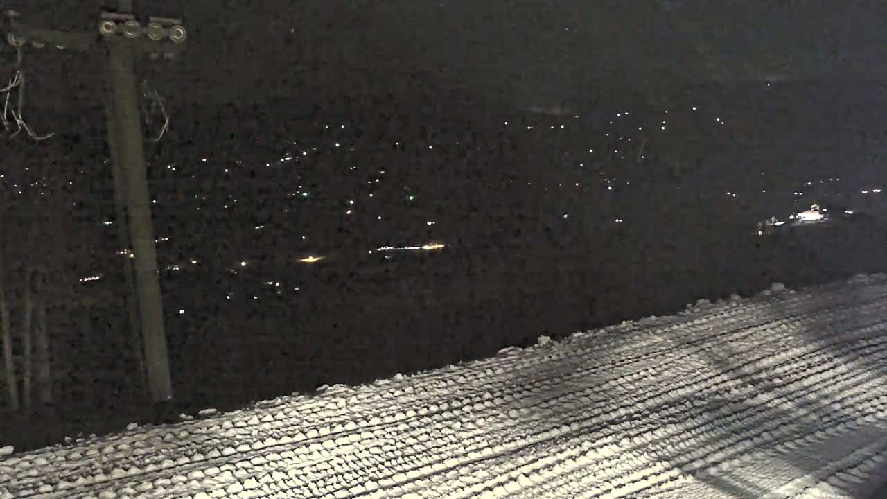 A snow-covered surface with tire tracks stretches toward a distant town illuminated by many small lights at night.