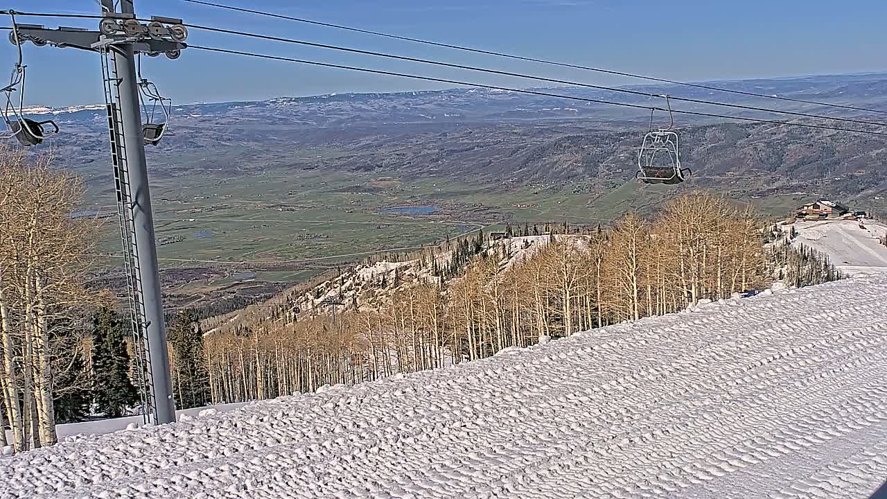 A ski lift chair ascends a snowy mountain slope overlooking a vast valley under a clear blue sky.