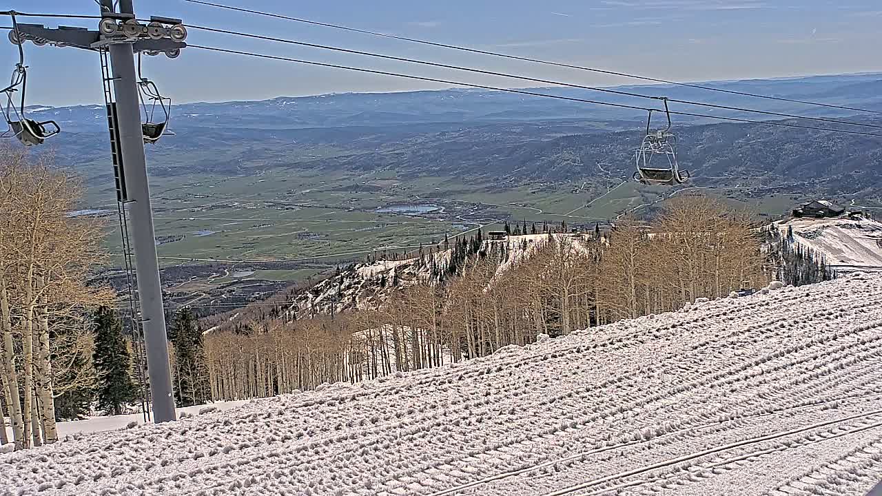 A ski lift chair ascends a snow-covered mountain slope overlooking a vast valley under a clear blue sky.