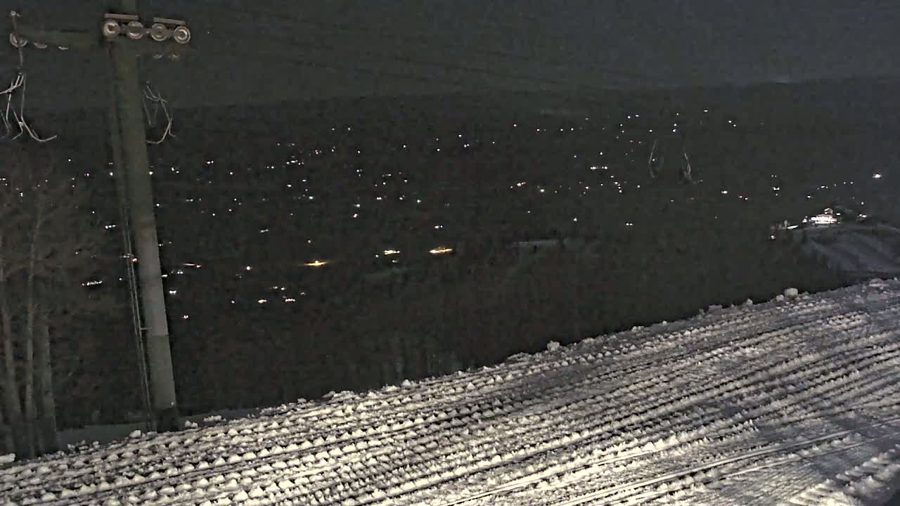 A snow-covered surface slopes downward toward a distant, dark valley illuminated by numerous small lights at night.