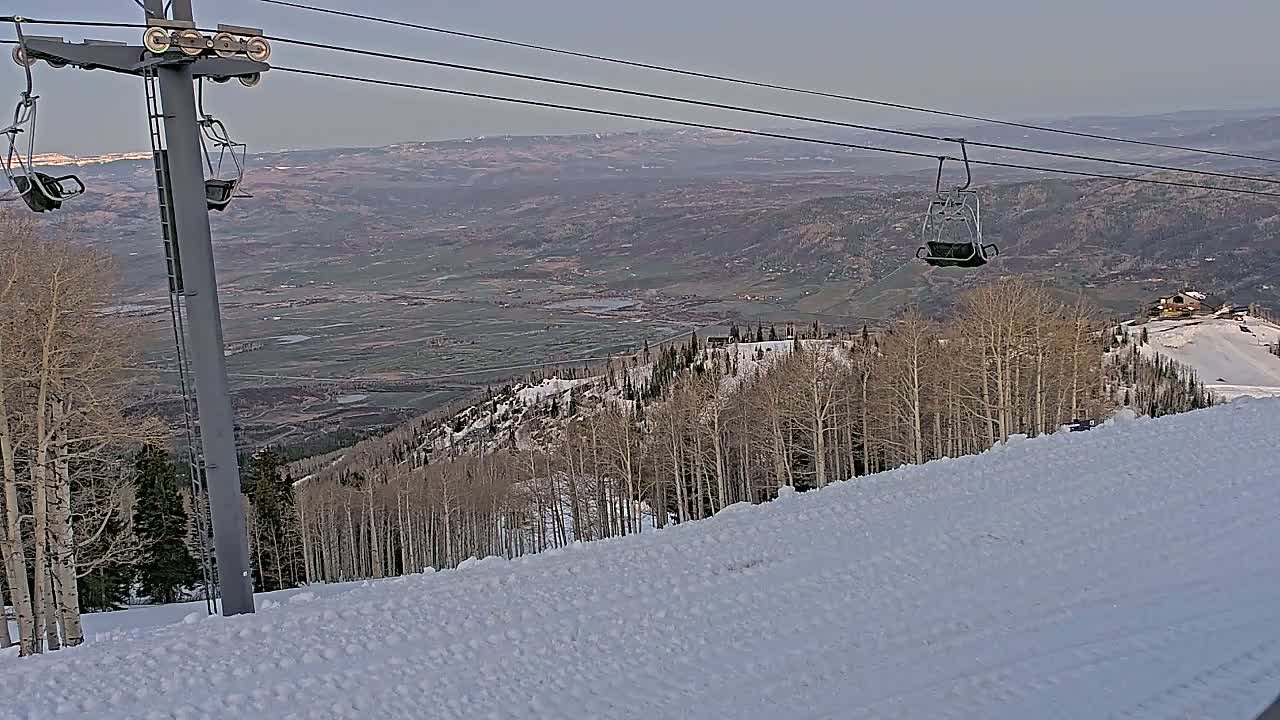 A snow-covered ski slope with a chairlift and a view of a valley under a hazy sky.