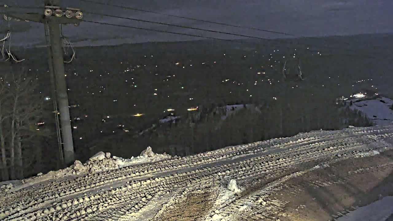 A snow-covered road with tire tracks winds down a hill at night overlooking a distant town speckled with lights.