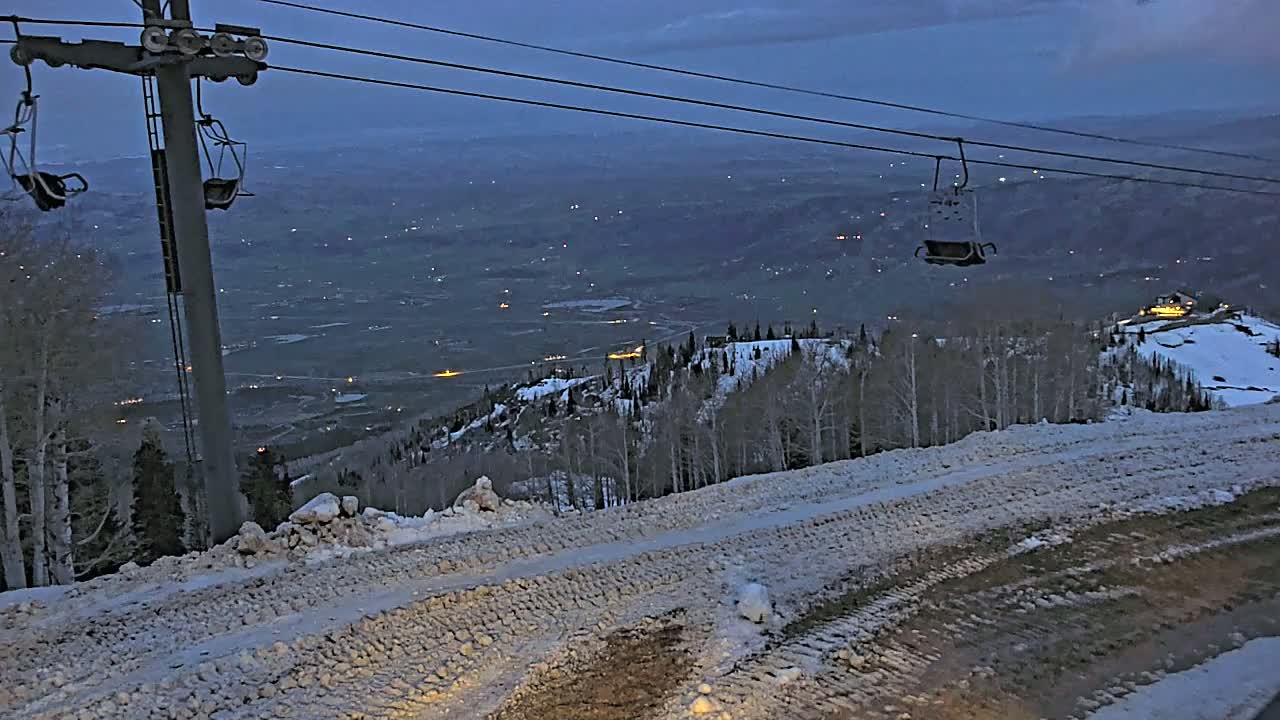 A ski lift at dusk overlooks a snow-covered mountainside and a valley speckled with lights below.