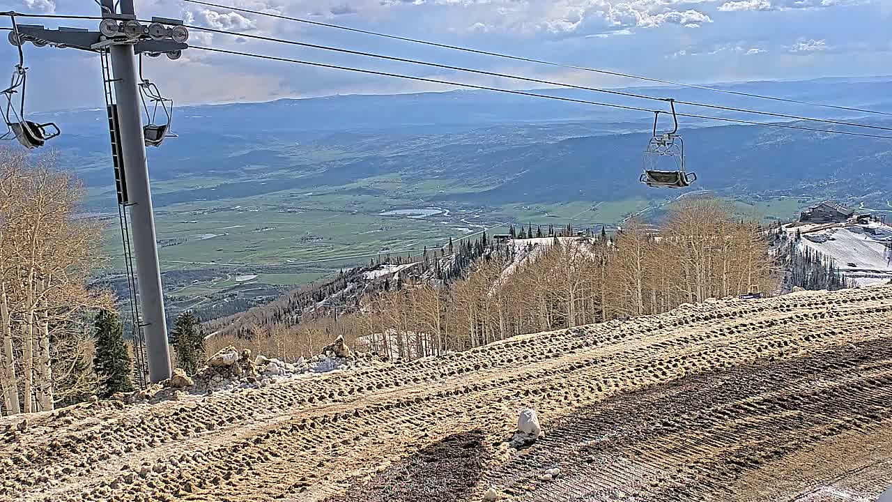 A mostly sunny day reveals a mountain vista with a chairlift, patchy snow, and tire tracks on a brown, dirt path.