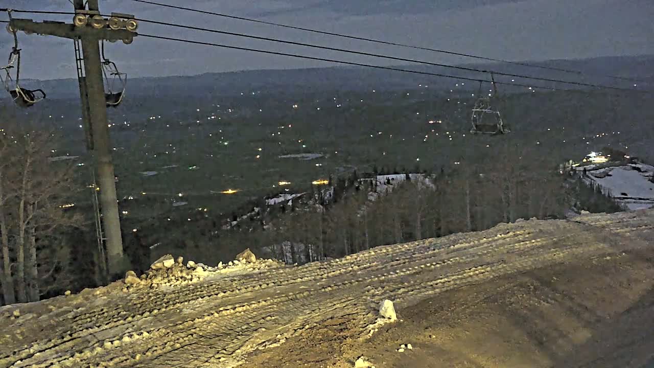 A ski lift at dusk overlooks a valley dotted with distant lights, with a snow-covered slope in the foreground showing tire tracks.