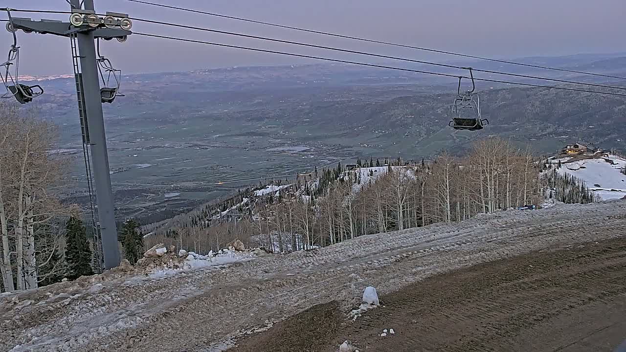 A ski lift with empty chairs overlooks a snow-dusted mountainside and a valley under a dusky sky.