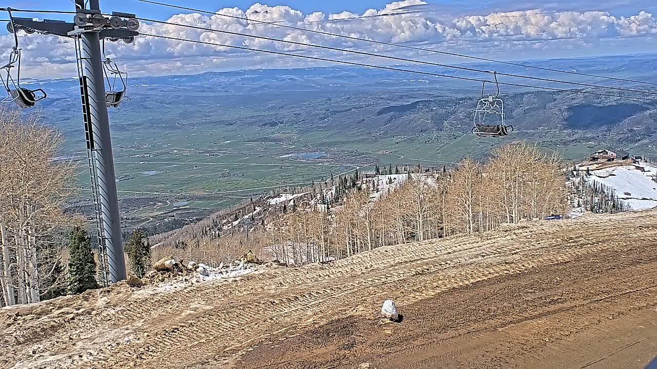 A chairlift ascends a mountainside on a partly cloudy day, overlooking a vast valley with patches of snow and bare trees.