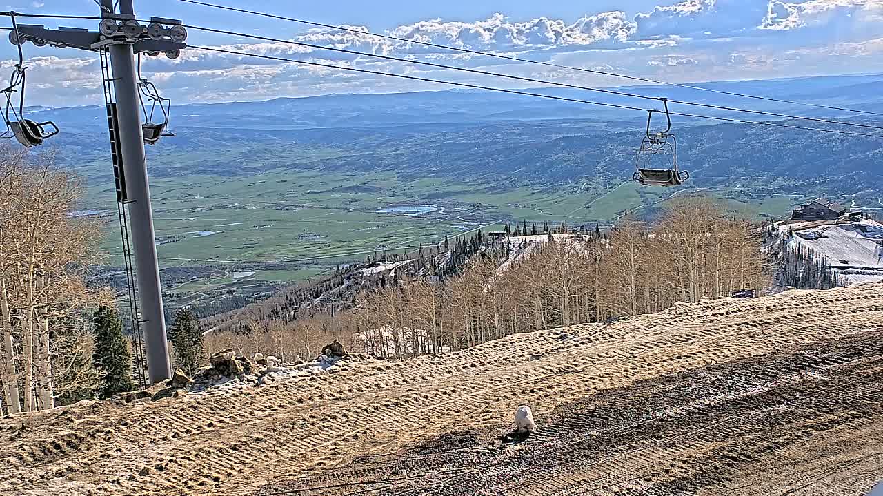 A mostly sunny day reveals a mountaintop ski lift overlooking a vast green valley, with patches of snow still visible on the mountainside and a groomed ski run in the foreground.