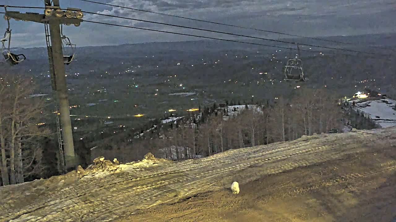 A ski lift overlooks a valley dotted with lights at night under a dark, cloudy sky.