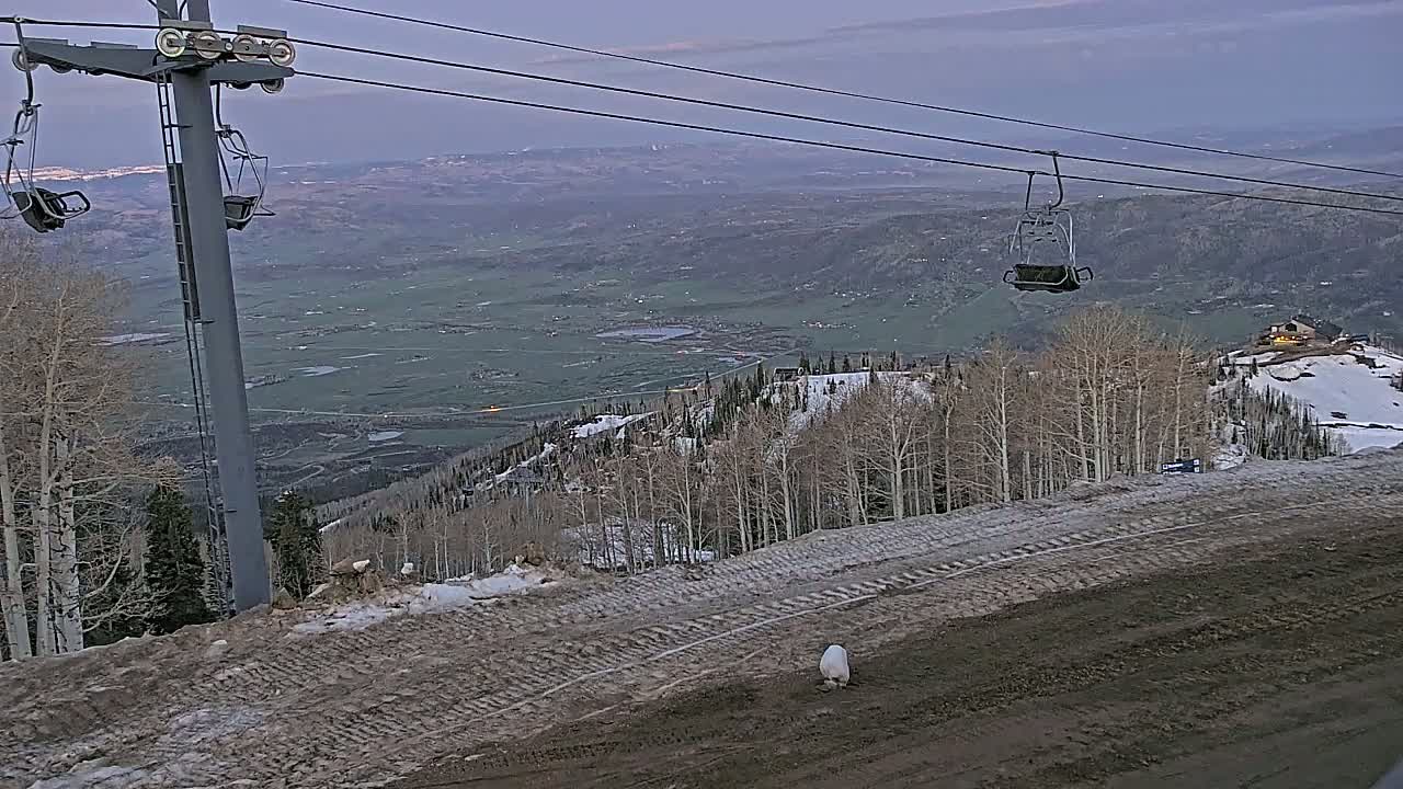 A ski lift with empty chairs overlooks a valley and snow-covered mountainside under a dusky sky.