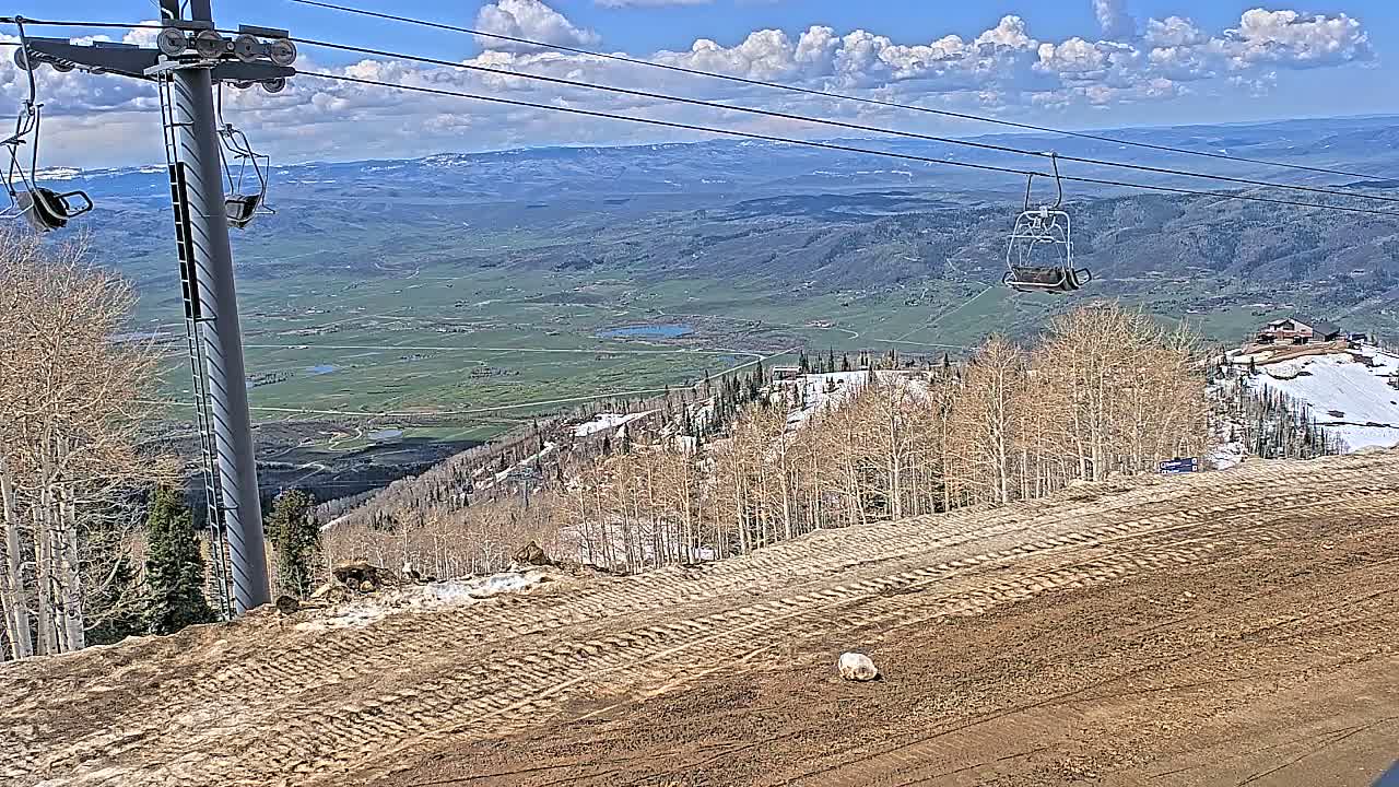 A mostly sunny day reveals a mountaintop ski lift overlooking a vast valley of green fields and distant mountains, with patches of snow visible on the slopes.