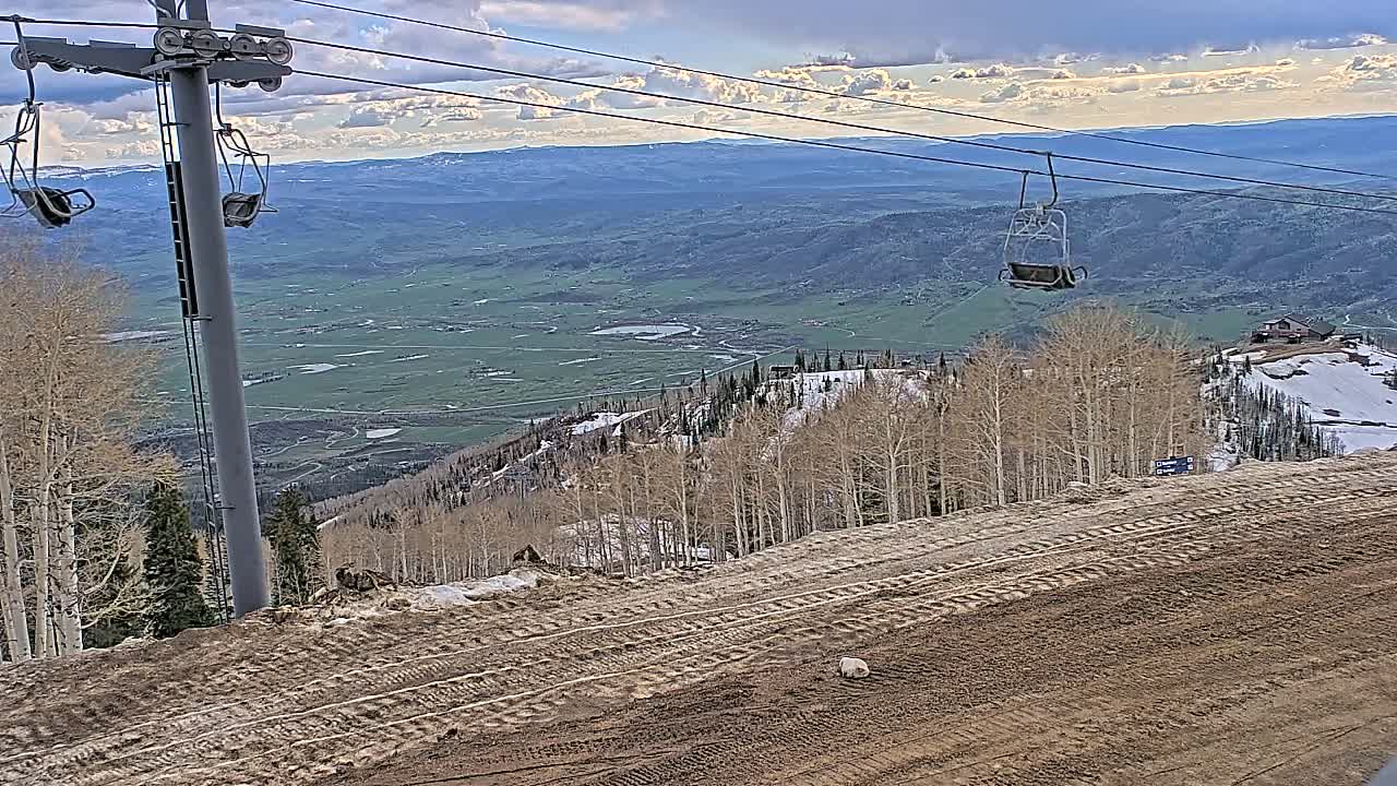 A ski lift chair hangs over a mostly barren mountain slope with tire tracks, overlooking a valley under a partly cloudy sky.