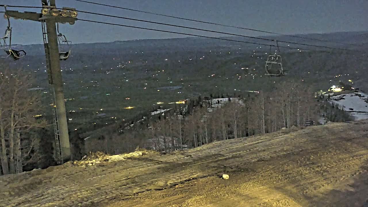 A chairlift at dusk overlooks a valley dotted with distant lights and sparsely wooded hillsides partially covered in snow.