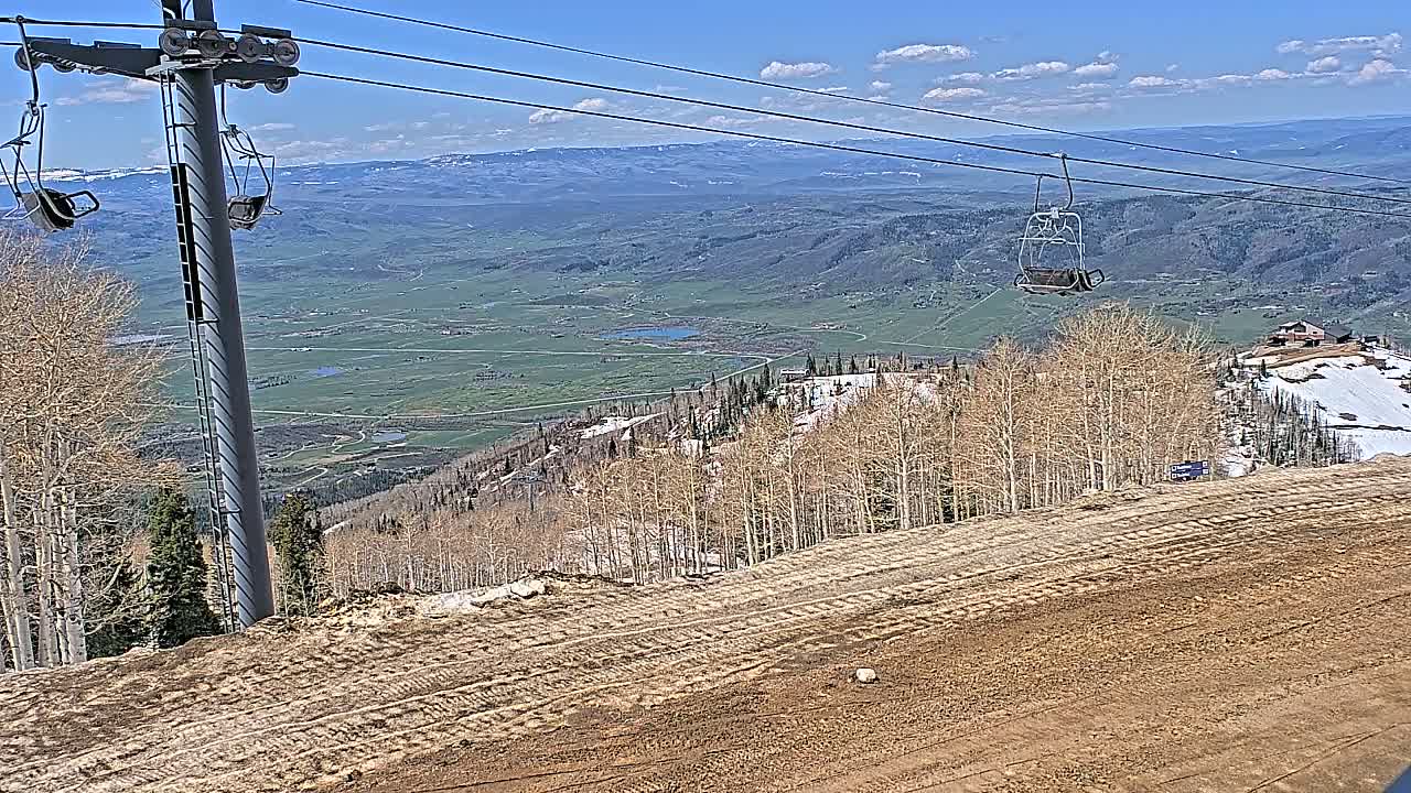 A ski lift chair ascends a mountainside on a sunny day with a clear view of a valley below, where patches of snow remain near the summit.
