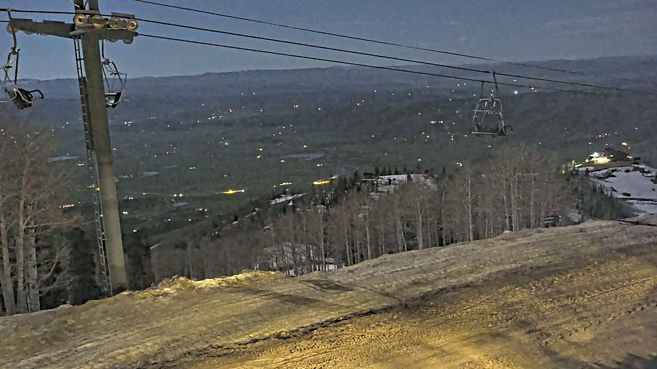 A ski lift chair and support structure overlook a valley speckled with distant lights at dusk or night.