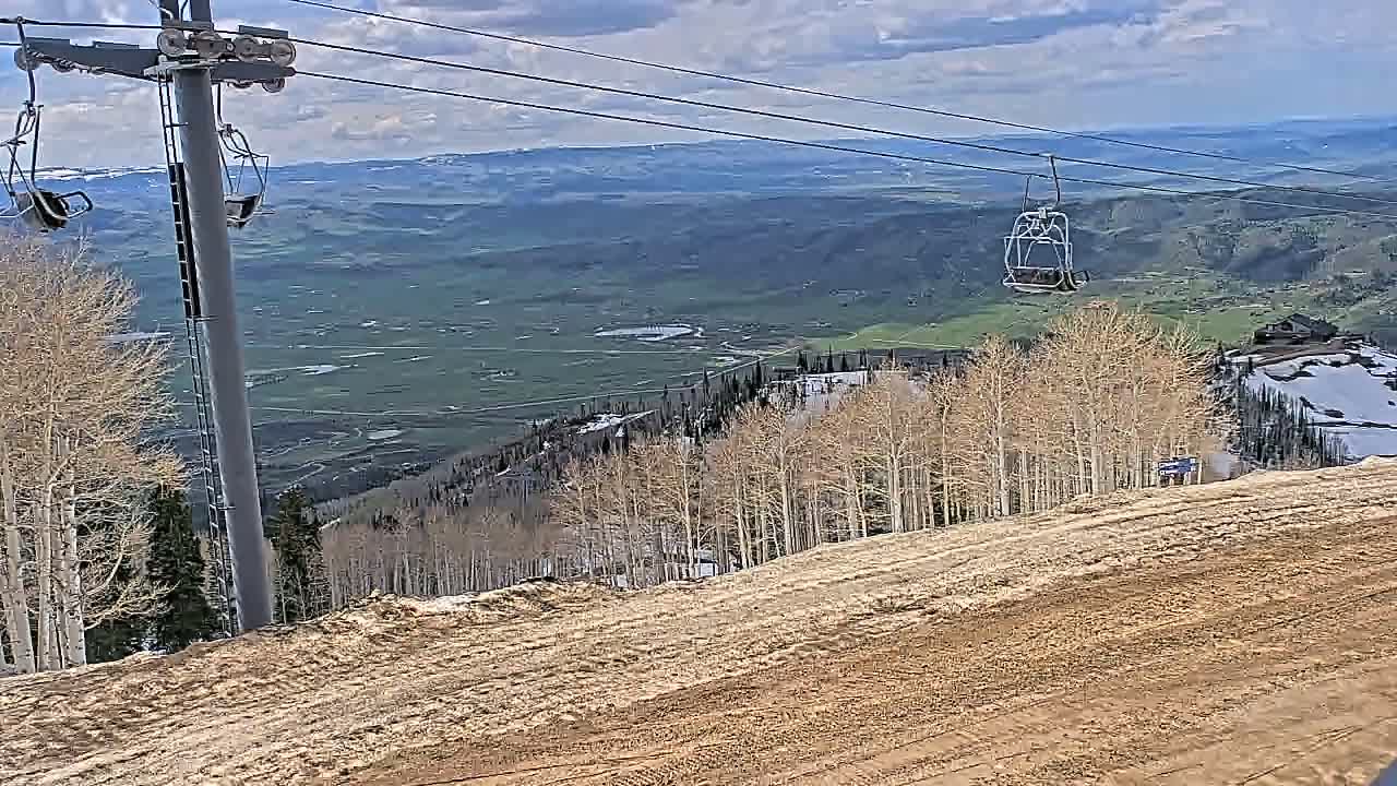 A chairlift ascends a mountain with bare aspen trees overlooking a valley under partly cloudy skies.