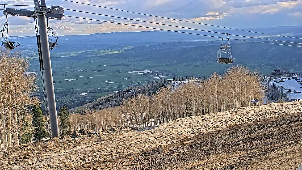A chairlift ascends a mountain slope overlooking a valley under a partly cloudy sky.