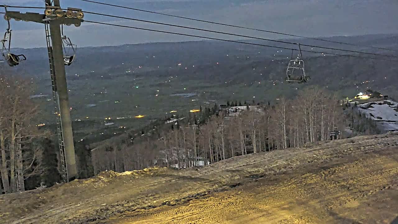 A ski lift with empty chairs overlooks a valley illuminated by scattered lights at dusk, with patches of snow visible on the ground.