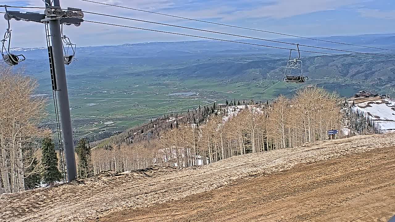 A ski lift chair ascends a mountainside with barren trees and patches of snow under a mostly sunny sky, overlooking a vast green valley.