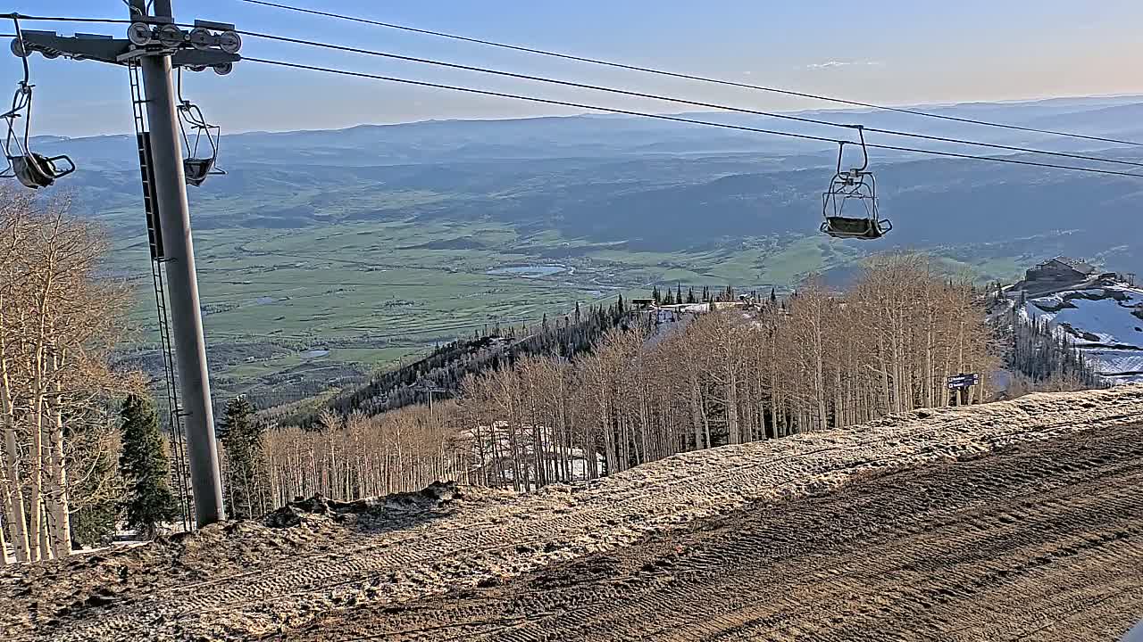 A ski lift with empty chairs ascends a mountainside overlooking a valley under a clear, sunny sky.