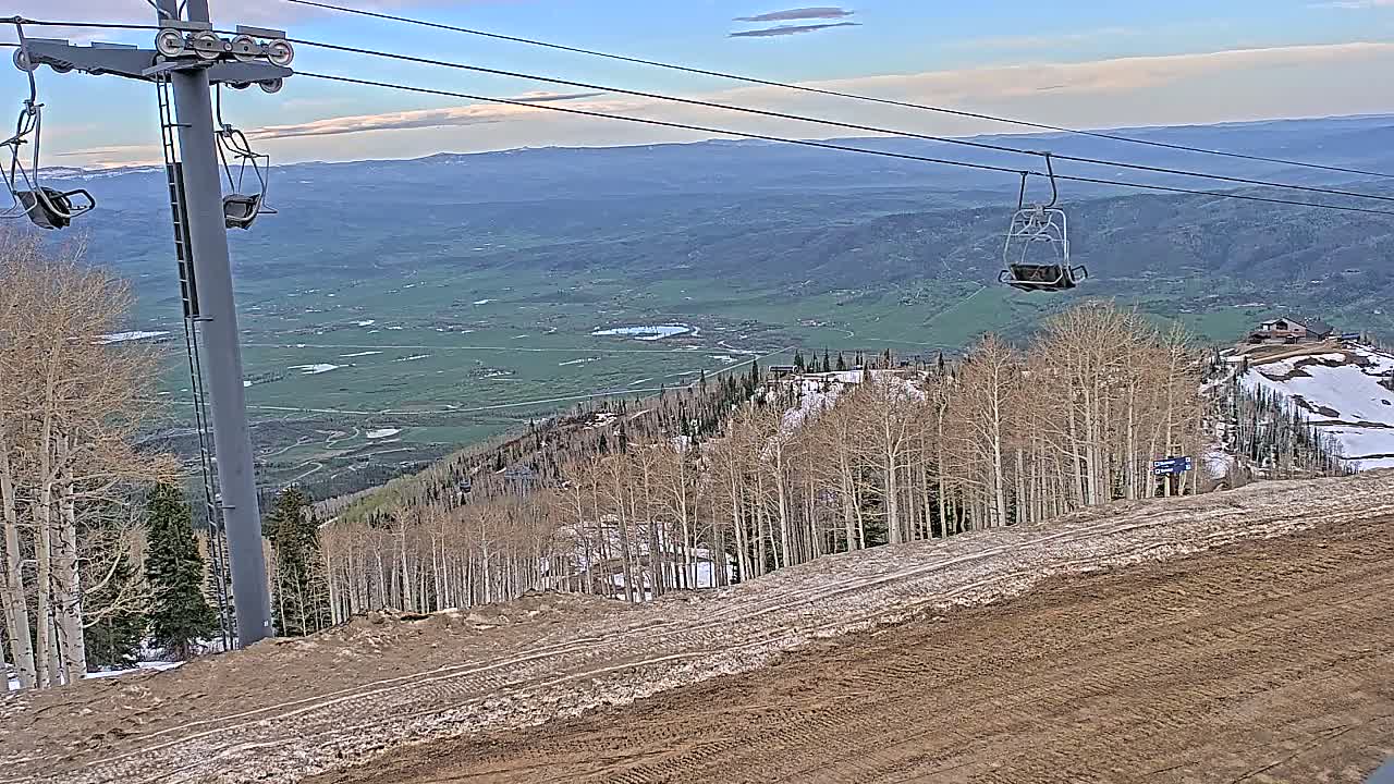 A ski lift chair ascends a mountainside overlooking a valley under a partly cloudy sky.